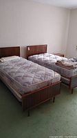 Two twin bed frames with mattresses positioned side by side in a room with light walls and green carpet.