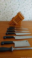 Six knives and sharpening rod arranged in front of wood block on tabletop