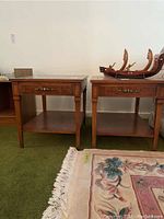 Pair of wooden end tables with brass hardware and lower shelves, on green carpet next to floral rug.