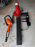 Top view of Black & Decker hedge trimmer, Toro leaf blower with nozzle attachment, and Mastercraft electric chainsaw laid on garage floor.