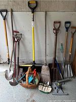 Full view of garden tools grouped against a wall, showing shovels, rakes, broom, hand tools in basket and roll, and sprinkler base on floor.
