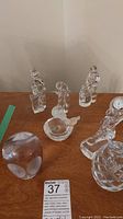 Six clear glass items on wooden surface: four figurines, one alien-face paperweight, one engraved faceted paperweight; one figurine has a clock mechanism.