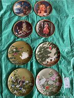 Eight decorative plates laid out on a green cloth showing four Norman Rockwell prints with people and four with bird and flower illustrations