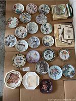 Full layout of 24 decorative china plates depicting various nature scenes, birds, flowers, and tiger, arranged on cardboard with certificates stacked at the top right.