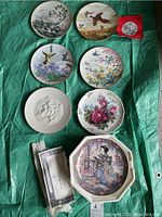Overview of all seven decorative china plates laid on green fabric, shows various bird, floral, embossed, and people figure designs. Certificate and original packaging for Lotus Blossom Maiden plate also visible.