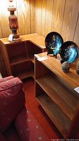 Photo showing corner shelf and bookshelf side by side with decorative plates and lamp on top.