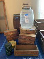 Assorted antique decor with tall clear glass jar and wooden crates labeled Sunshine Farms and Meadow Gold, arranged on a blue floor covering.