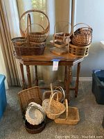 Photo showing multiple baskets arranged on a wooden table and on the floor beneath it. Varied shapes and sizes visible, some round, some rectangular, all wicker or wood tone.