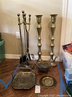 Photo of brass fireplace tools with stand, pair of tall candleholders, octagonal brass tray, and decorative vintage Mexican plate on floor.