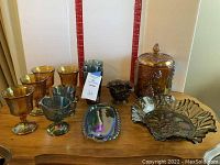 Photo showing assorted iridescent carnival glass serving pieces including goblets, bowls, a covered jar with lid, and serving trays arranged on wooden table.