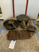 Overall view of assorted cast iron cookware and tools including pans, lid, spatula, and iron on carpet.