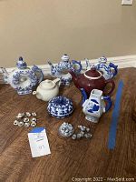 Photo showing five medium-sized ceramic teapots and decanters in blue and white floral designs plus one dark red teapot, small cups, and a lidded bowl.