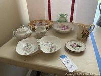 Assorted china pieces arranged on a table, including divided dish, sugar bowl, creamer, small plates, pastel green pitcher, and white bowl with floral decorations.