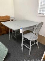Photo shows gray painted wooden dining table with two matching spindle back chairs. One chair clearly shows a split seat.