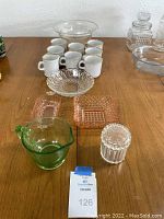Photo showing arranged items on table: large clear ribbed glass bowl, white ceramic mugs, pink depression glass dishes, green vintage measuring cup, clear ribbed canister, additional clear glass bowls.