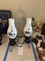 Two matching vintage-style lanterns with white glass shades featuring black floral designs alongside a silver metal decorative bird cage, displayed on a flat surface.