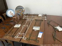 Wide view of six metal plate display hangers laid out on a wooden table with two decorative plates on stands at one end.