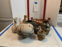 Photo showing group of pottery pieces, copper-colored metal dish, and wooden bird figurine arranged together on white surface with red measuring scale in background.