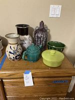 Photo showing six stoneware and pottery decorative items arranged on wooden furniture including various vases, a teapot, and a lidded dish.