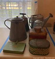 Vintage metal cowboy coffee pots of various sizes with handles and lids, alongside assorted vintage metal tins mostly rectangular or oval in shape, placed on a white surface near a window with sheer white curtains.