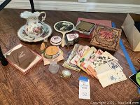 Full view of the vintage vanity accessories lot laid on wooden floor showing floral pitcher, handkerchiefs, compact mirrors, jewelry containers, and music box.