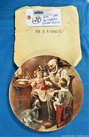 Plate with original backing paper/story, displaying the artwork "The Toy Maker" by Norman Rockwell. The paper backing identifies it as a 1977 edition by Knowles Fine China, numbered 6924 A. The plate shows the image of an old man making toys, two children watching, and a dog nearby.