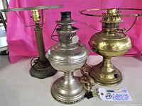 Full view of three vintage lamps: silver Aladdin oil lamp front and center, brass lamp to right, metal electric lamp to left