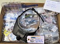 Box lot with jewelry making seed beads in resealable bags and black adjustable head magnifier positioned on top.