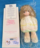 Doll in original white box, sealed in plastic, showing the long curly blonde hair and satin off-white dress with yellow, pink, and green trim.