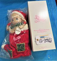 Photo showing the Precious Moments Christmas Eve doll sealed in plastic with original box standing upright next to it. Doll dressed in red velvet stocking with holly patches, visible PM tag attached.