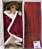 Full view of the doll in the box, showing red velvet cloak with white faux fur trim. Wood-grain print box with Dynasty Doll Collection branding.