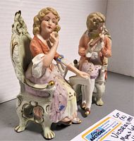 Front view of both Victorian porcelain figurines seated in ornate chairs, showing lady holding handbag and man playing flute