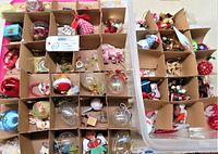 Top view of a compartmentalized red storage box filled with various vintage Christmas ornaments including glass baubles, ceramic figures, cloth decorations, and metal charms.