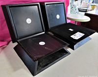 Four black wood display stands with square top platforms, matte finish, with white round stickers visible, arranged together showing top and angled side views, accompanied by a white price tag.