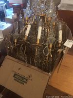 Antique crystal chandelier in a medium cardboard box showing multiple candle-like bulbs and crystal components packed for storage or transport.