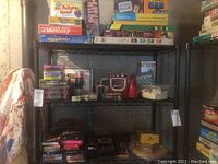 View of metal shelving unit with multiple board games stacked including Memory, Mr. Potato Head Playful Pictures, Spy Web, and other boxed games.