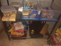 Plastic bins with assorted game pieces, tokens, and cards along with stacked board games and wooden trays on metal shelving.