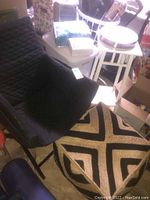 Photo of black quilted armchair, woven foot stool with geometric design, white laundry basket with lid, white wooden side table, other dorm essentials visible