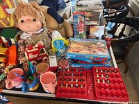 Wide view of various vintage collectible toys including a plush doll with overalls, a blue plush toy, boxed Fischer Technik construction sets, a tray of Mighty Beanz, and vintage cups with utensils arranged on a table.