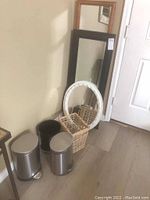 Two framed mirrors stacked against the wall, one light wood frame and one dark wood frame, with a white oval wicker basket and three metal waste baskets in front.