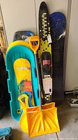 Full lot showing two snowboards leaning against wall, two sleds stacked on the floor, and two yellow snow shovels.