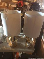 Two brass base table lamps with white cylindrical shades on a white table in cluttered basement setting.