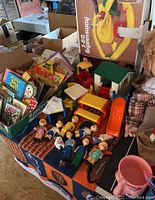 Lot overview showing vintage children's books standing in boxes, wooden toy figures lying on a blanket, Little Tykes dollhouse furniture parts, and the boxed I-80 Expressway toy set in background.
