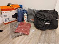 The complete lot arranged on floor showing all items including air bed boxed with label, two fleece blankets, Camelbak water bottle, gray Kenmark ski bag, black Voyager saddle bag, travel neck pillow, and magic bag