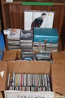 Photo showing large cardboard box filled with assorted music CDs in jewel cases, cassette carrying case, stacks of CDs on wood cabinet shelf, and Michael Buble Christmas vinyl album propped against back wall.