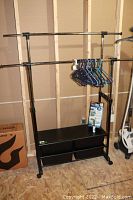 Full view of black metal and plastic adjustable clothing rack with storage shelves, several plastic hangers hanging on upper bars, Space Bag vacuum storage bag package and lint roller on shelf below, placed in unfinished room.