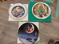 Three Christmas-themed collector plates in original boxes shown on wooden floor. Plates feature Santa Claus and Christmas scenes.