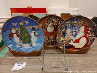 Set of four Christmas themed collector plates displayed on wooden stands; plates show snowman with Christmas tree, Santa Claus scenes; vibrant holiday colors visible