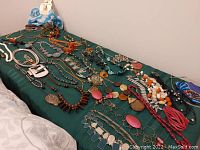 Wide view of a collection of costume jewelry necklaces laid out on a dark green cloth, showing a variety of colors and styles including beaded and pendant necklaces