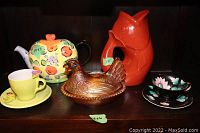 Full view of lot on dark shelf showing 5 items including covered glass chicken bowl, 2 teacups with saucers, Linda Miller teapot, red fish decanter